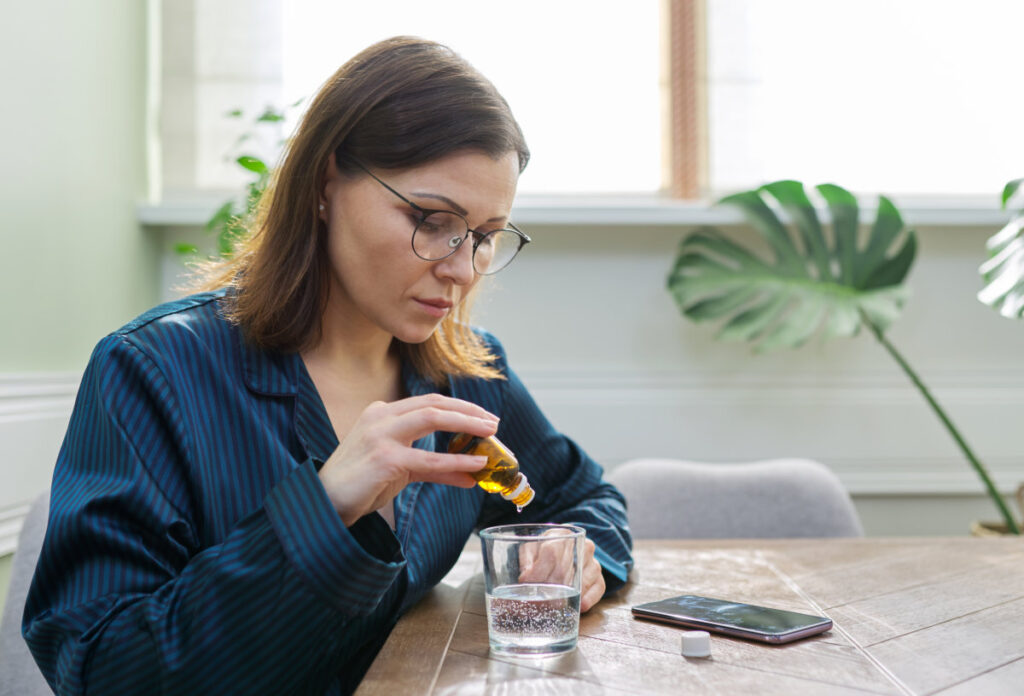 Nervous disorder, depression, nervousness, age-related health problems. Mature woman dripping drops with glass of water, drinking sedative medicine, at home in pajamas, copy space